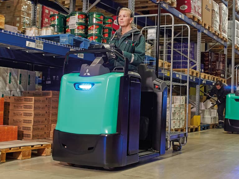 Photographie mettant en scène une cariste au volant d'un chariot préparateur de commandes Mitsubishi Forklift, circulant avec aisance à l'intérieur d'une plateforme logistique